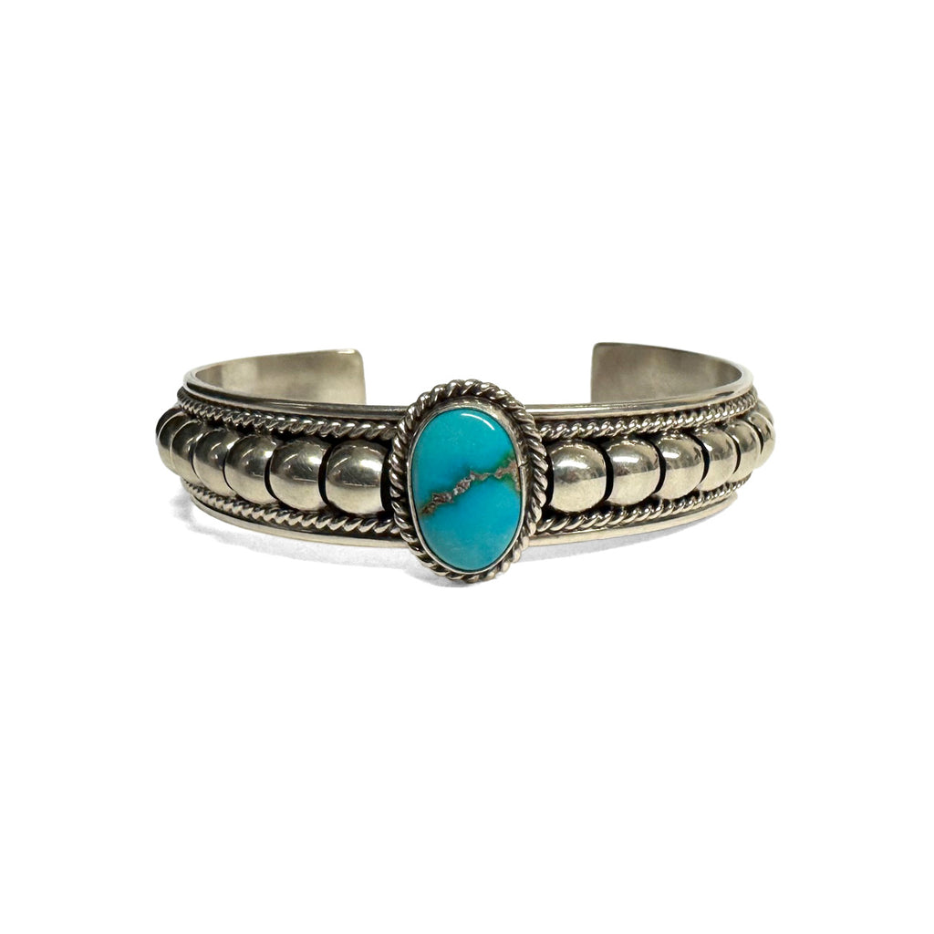 Front view of a silver bracelet featuring artistic accents and a turquoise stone at the center. Turquoise stone has a natural vein running through the center. Bracelet is on a white background.