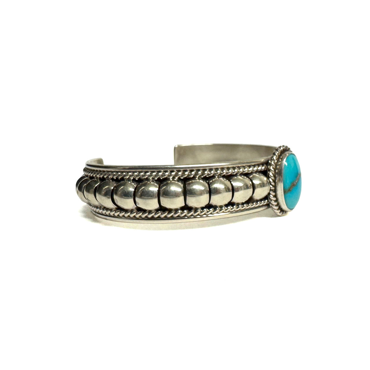 Side view of a silver bracelet featuring artistic accents and a turquoise stone at the center (facing right). Turquoise stone has a natural vein running through the center. Bracelet is on a white background.