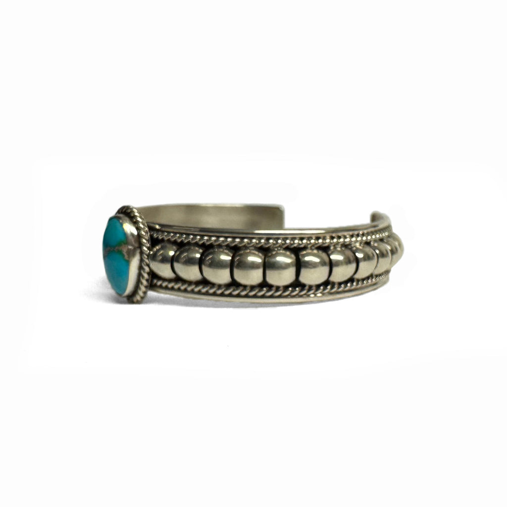Side view of a silver bracelet featuring artistic accents and a turquoise stone at the center (facing left). Turquoise stone has a natural vein running through the center. Bracelet is on a white background.