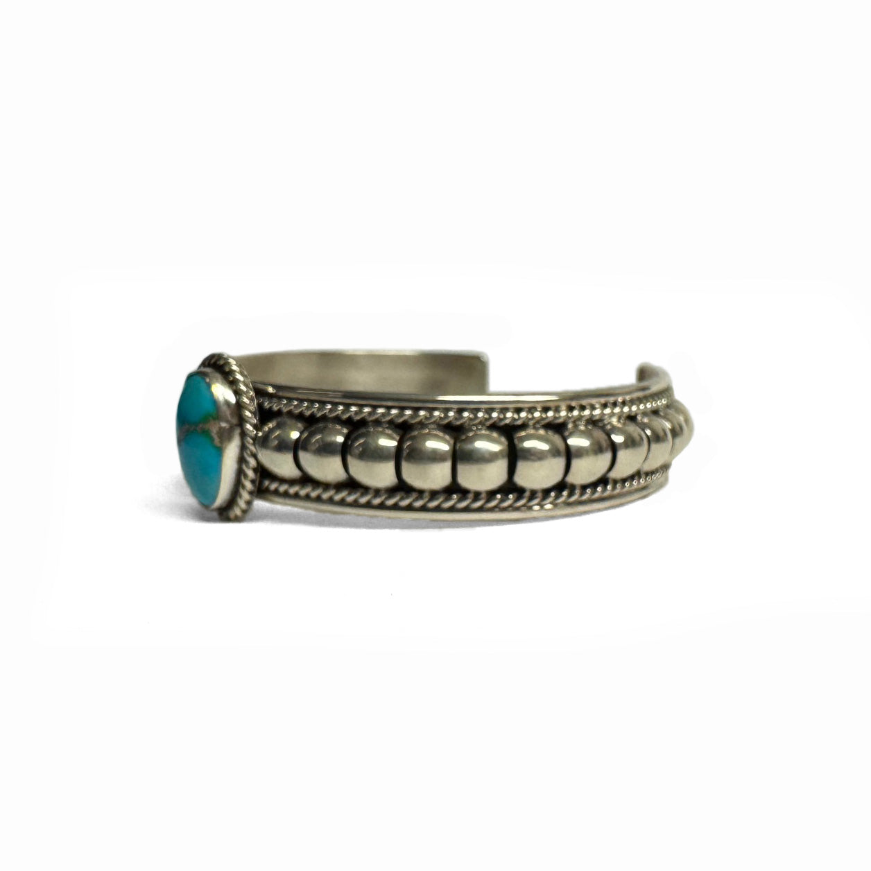 Side view of a silver bracelet featuring artistic accents and a turquoise stone at the center (facing left). Turquoise stone has a natural vein running through the center. Bracelet is on a white background.