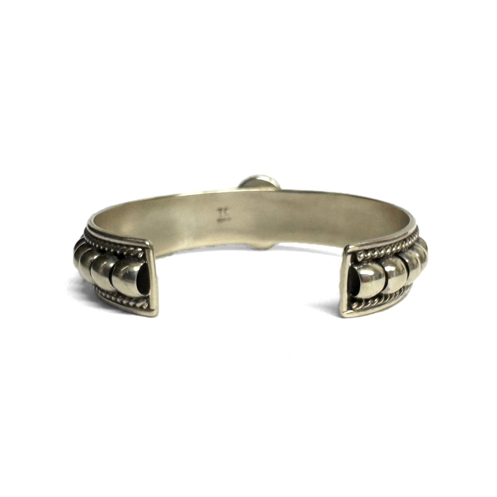 Back (inside) view of a silver bracelet featuring artistic accents and a turquoise stone at the center (facing away and not visible aside for silver setting). Bracelet is on a white background.