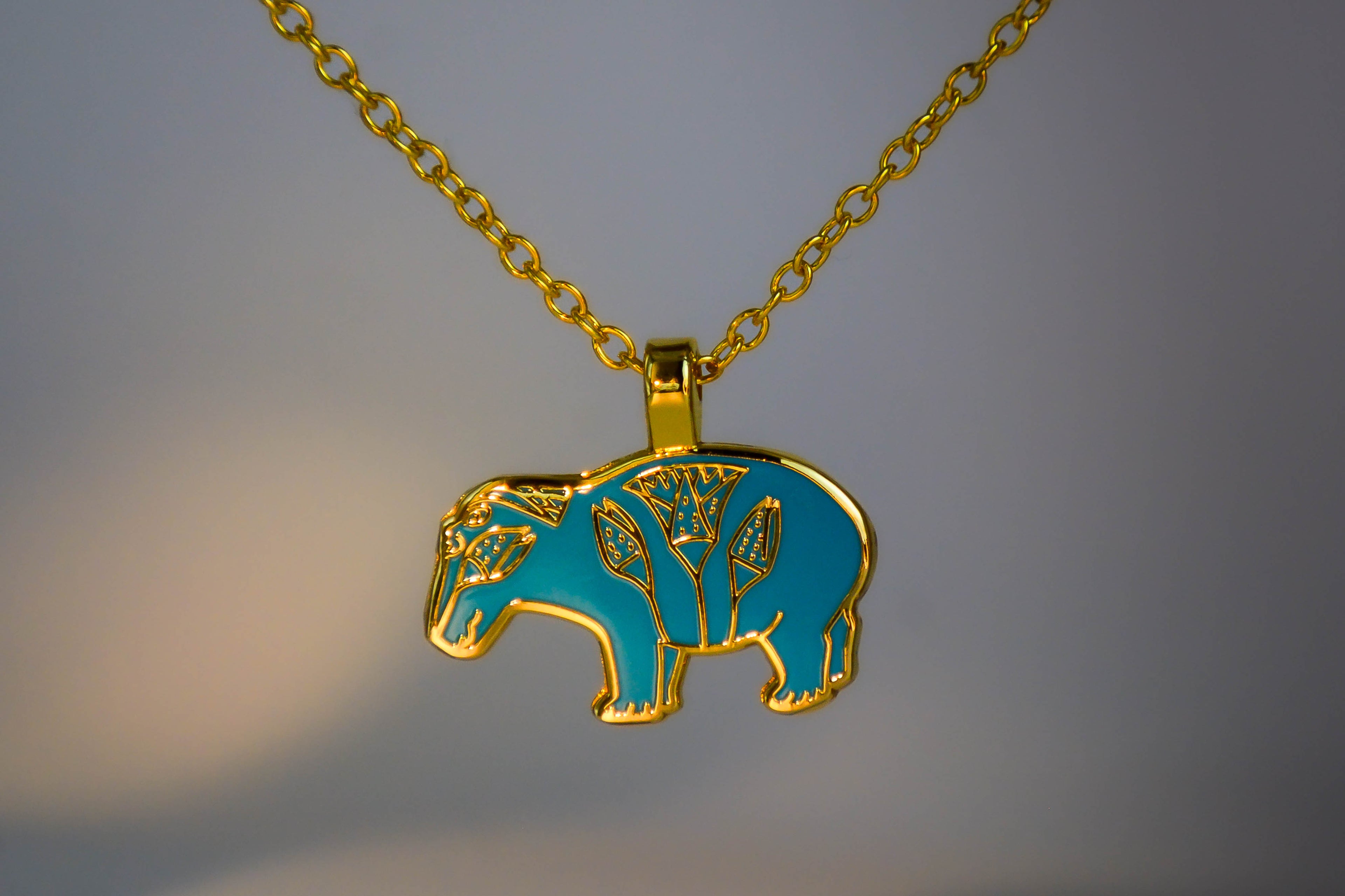 Enamel hippo necklace on off-white with purple, blue, and orange toned lighting background.