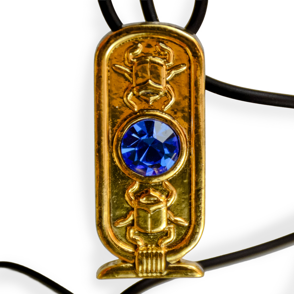 Cartouche-shaped ovular gold pendant with blue jewel in the center, a golden scarab beetle on the top and bottom of the jewel. Pendant on black necklace with white background. 