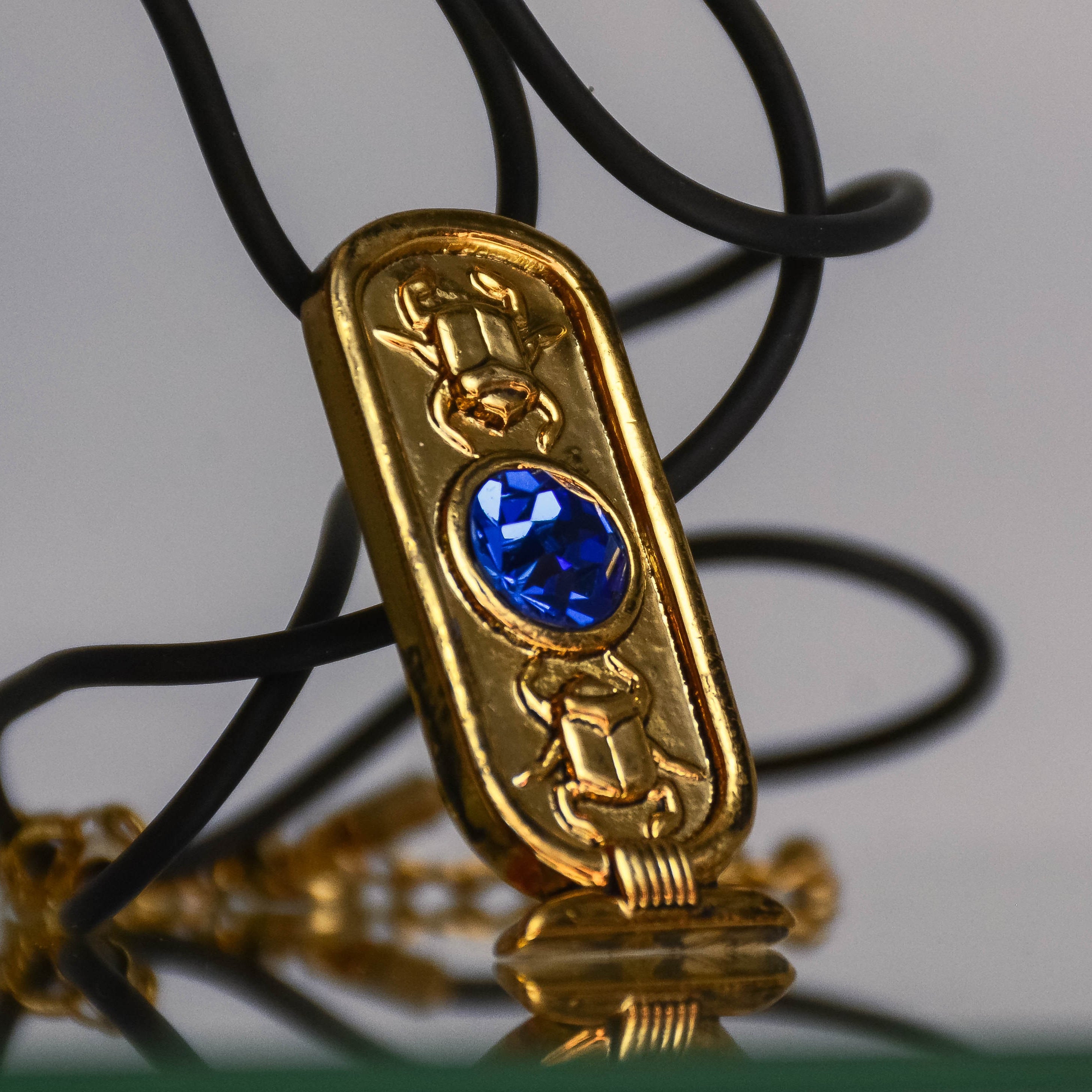 Gold Pendant is shown at a 45-degree angled view and is reflected below itself elegantly. Pendant on a black necklace with a gold clasp and chain, on an off-white background. 